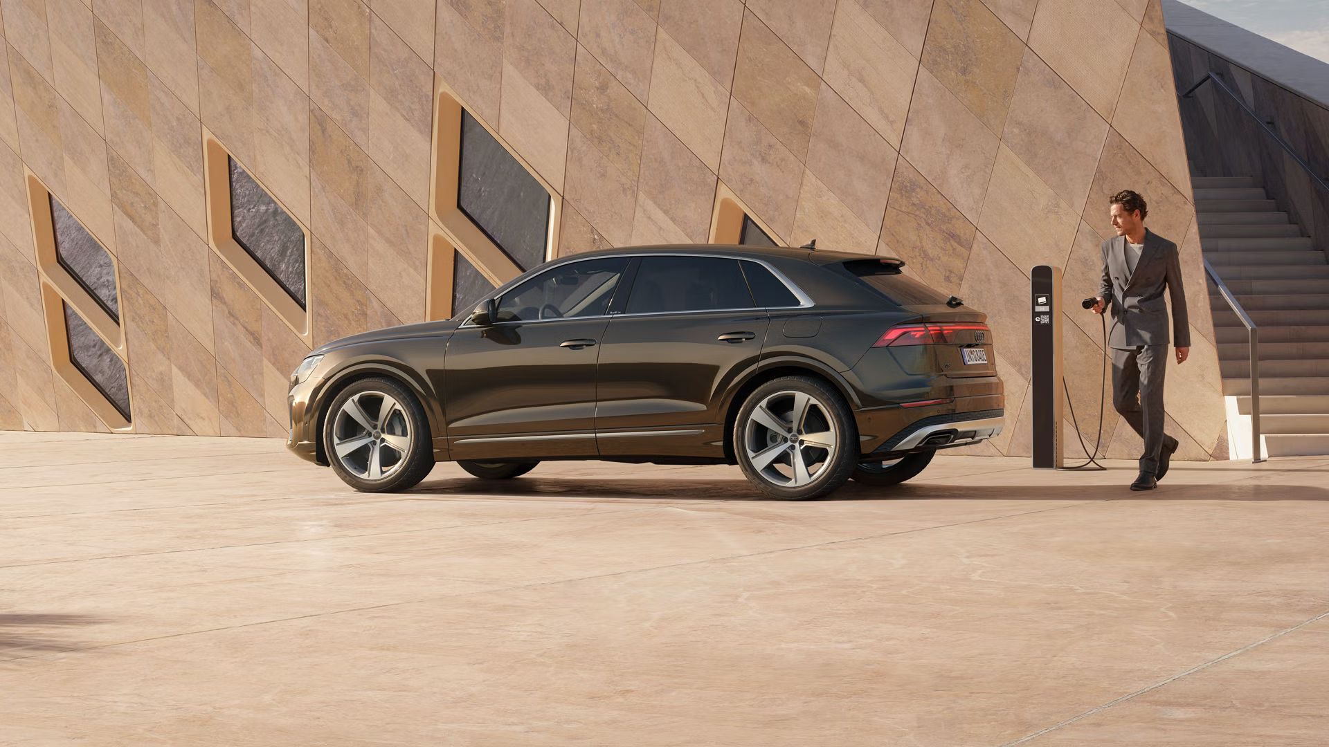 Der Q8 SUV e-hybrid schräg von der Seite parkt vor einem futuristischen Gebäude. Ein modisch gekleideter junger Mann steht hinter dem Fahrzeug und hält den Stecker einer Ladesäule in der Hand.