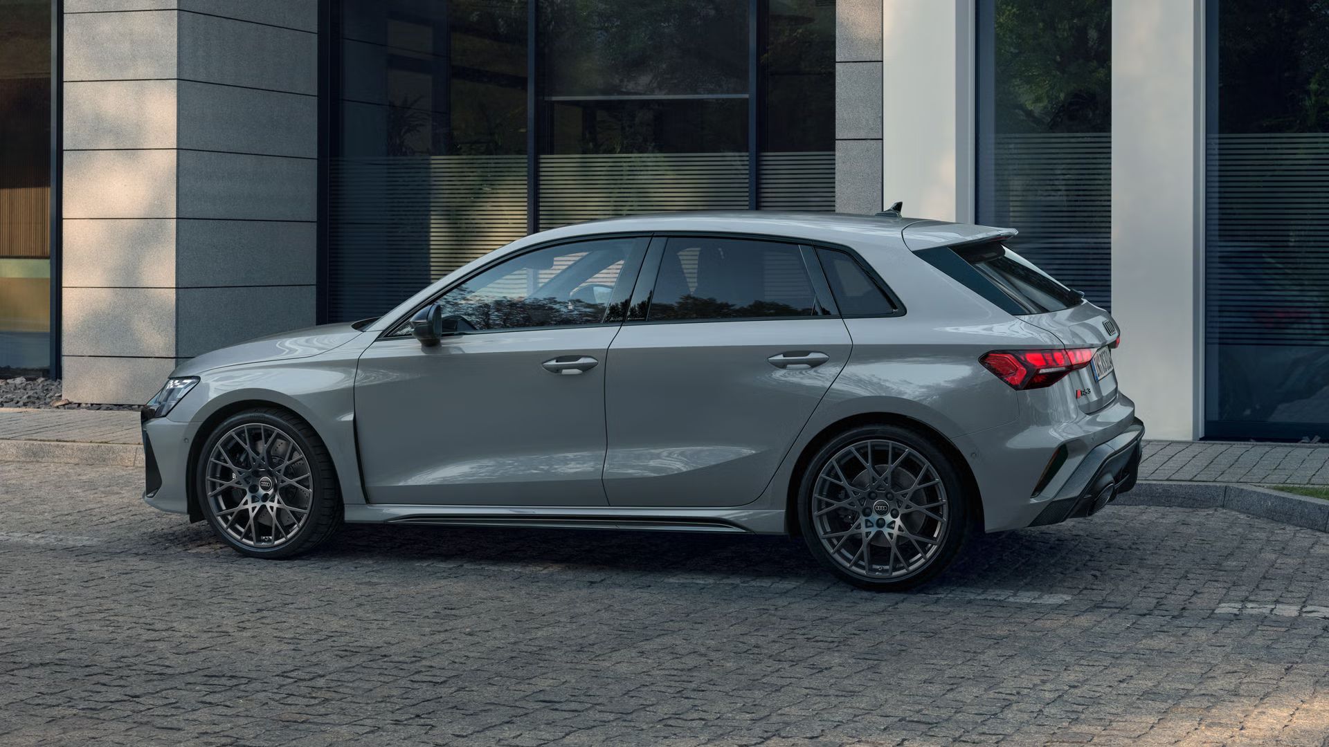 Der RS 3 Sportback mit Indivuduallackierung in Pfeilgrau Perleffekt von der Seite parkend auf Kopfsteinpflaster, im Hintergrund Umrisse eines modernen Gebäudes.
