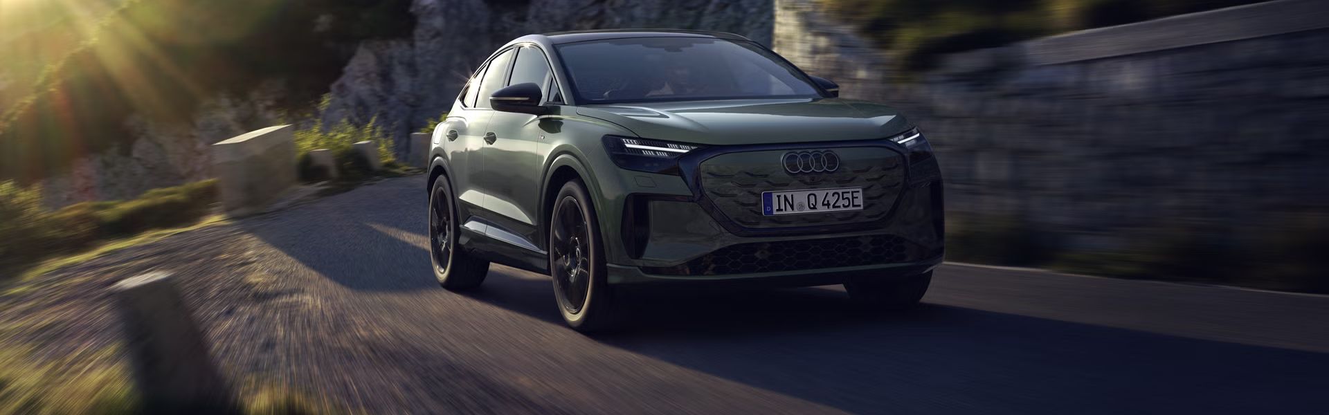 Der Q4 Sportback e-tron bei der Fahrt auf einer Bergstraße. Sonnenstrahlen von links oben beleuchten das Bild, das Fahrzeug wirft Schatten nach vorne.